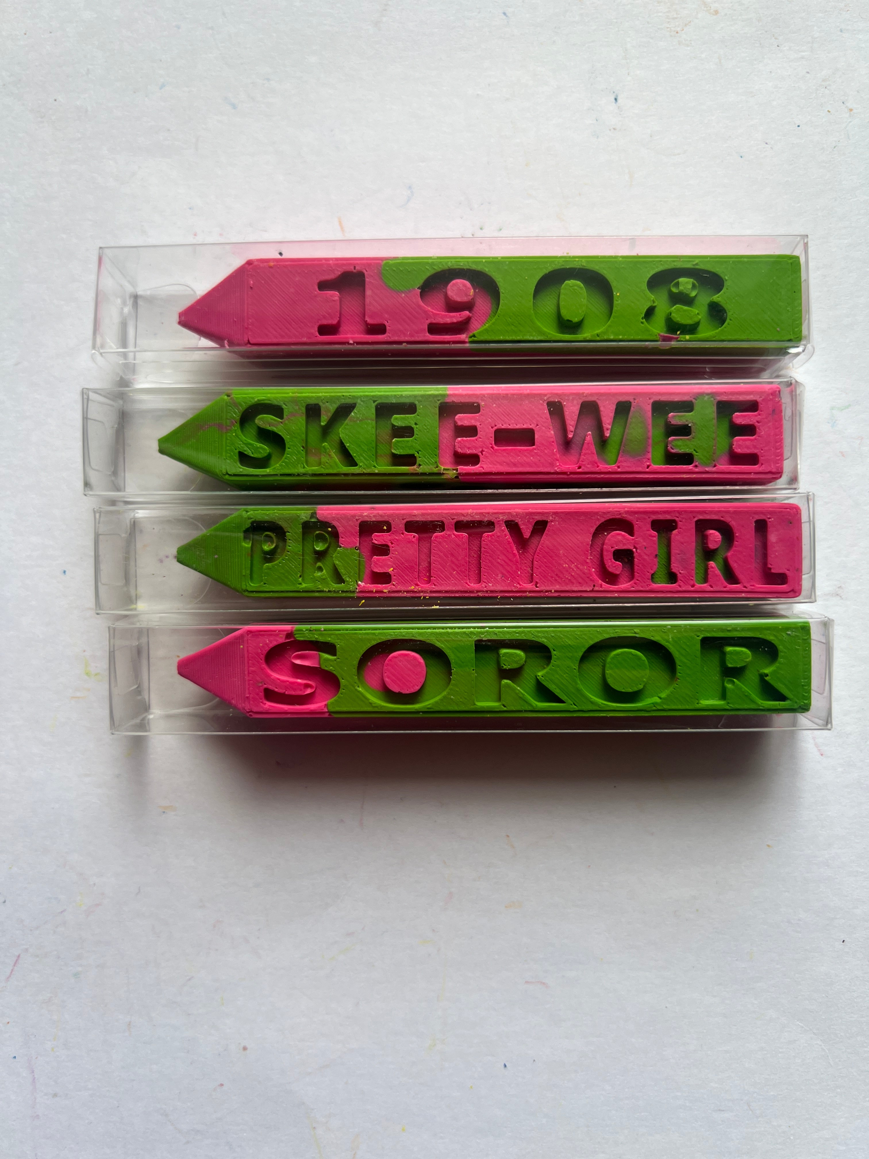 Four novelty crayons in clear plastic packaging, featuring pink and green crayons with embossed names and numbers: '1908', 'SKEE-WEE', 'PRETTY GIRL', and 'SOROR' on a white background.