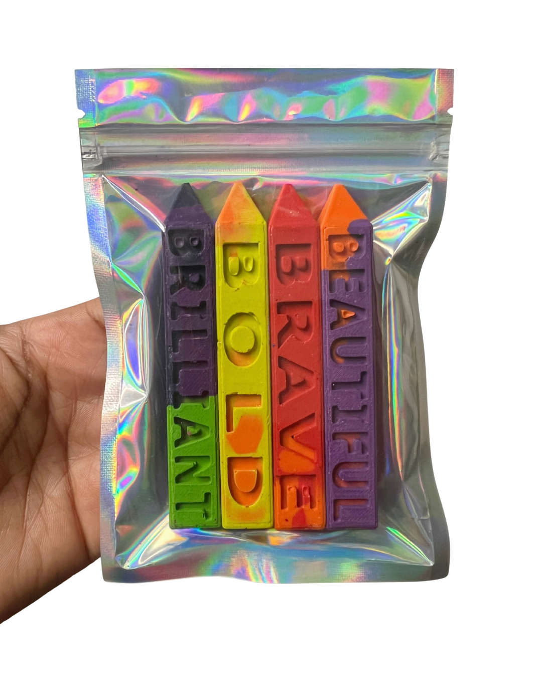 A hand holding a clear holographic mylar pouch containing four crayon-shaped items with engraved words: BRILLIANT, BOLD, BRAVE, and BEAUTIFUL, shown against a plain white background.