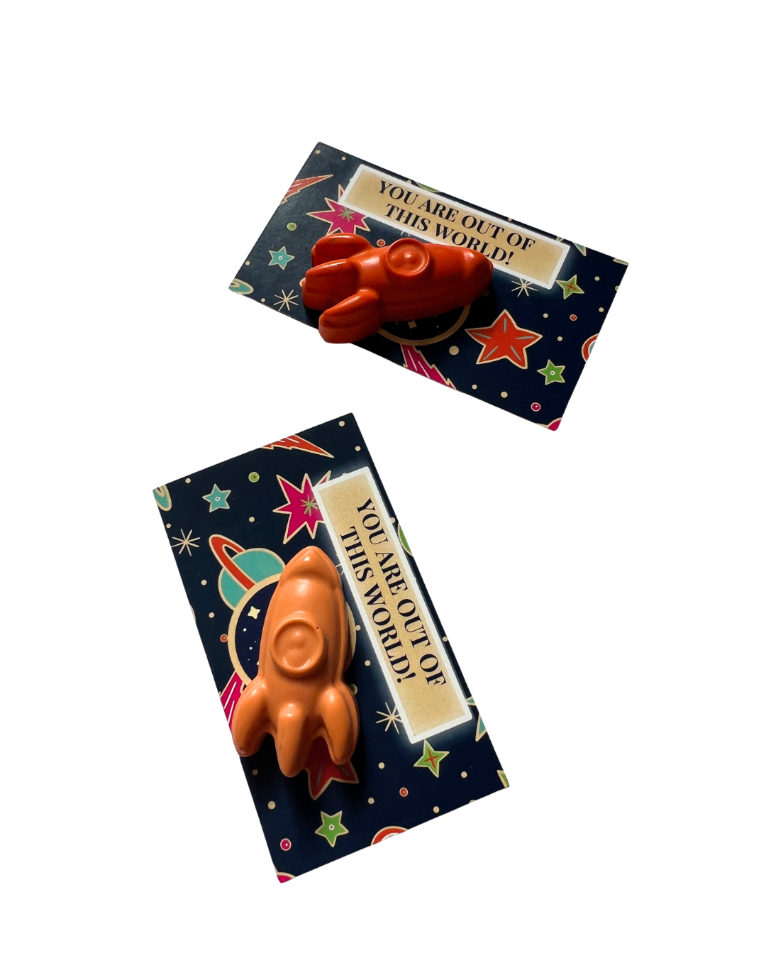Two small rocket-shaped crayons displayed on dark printed cards with a space-themed design and the phrase 'YOU ARE OUT OF THIS WORLD!'.