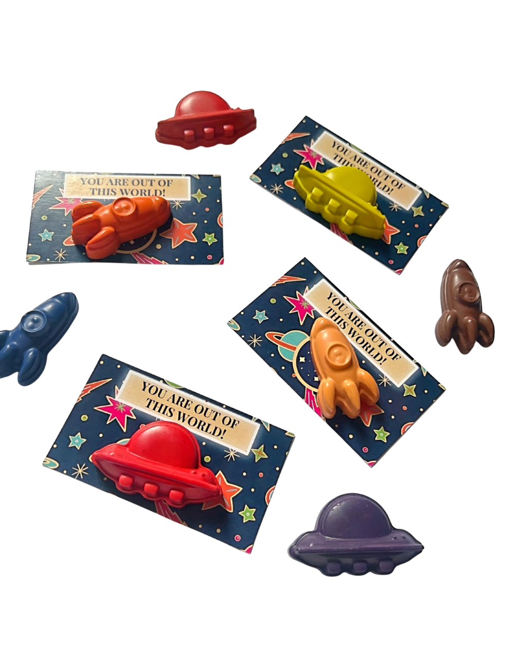 Seven small space-themed crayons shaped like rockets and spaceships arranged on a plain white background with their printed backing cards visible.