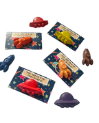 Seven small space-themed crayons shaped like rockets and spaceships arranged on a plain white background with their printed backing cards visible.