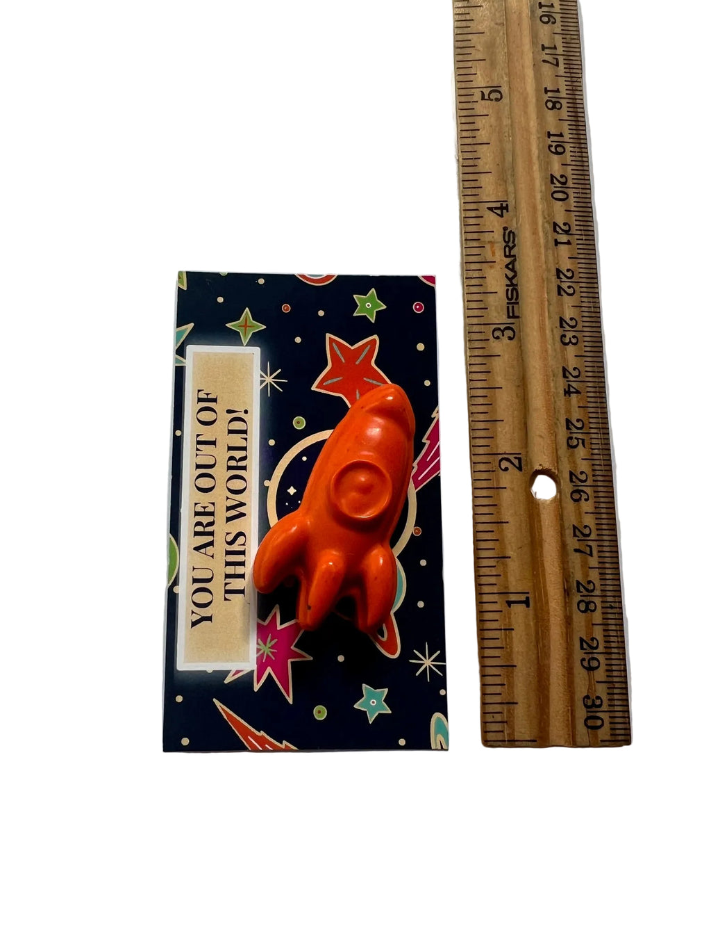 An orange rocket crayon displayed on a dark blue space-themed backing card next to a wooden ruler for scale, on a white surface.