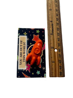 An orange rocket crayon displayed on a dark blue space-themed backing card next to a wooden ruler for scale, on a white surface.