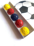 Five soccer-themed crayons in yellow, red, and navy blue, arranged inside a long white box next to a large soccer ball graphic on a white background.